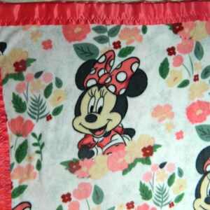 May include: A white baby blanket featuring Minnie Mouse with a red polka dot bow and dress, surrounded by floral patterns in pink, yellow, and green. The blanket has a red satin border.