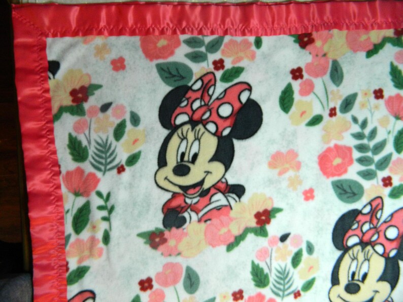 May include: A white baby blanket featuring Minnie Mouse with a red polka dot bow and dress, surrounded by floral patterns in pink, yellow, and green. The blanket has a red satin border.