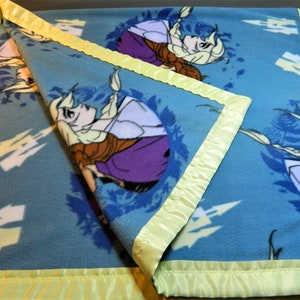 May include: A blue fleece blanket with a yellow satin border. The blanket features a repeated pattern of a cartoon image of a girl with long blonde hair.