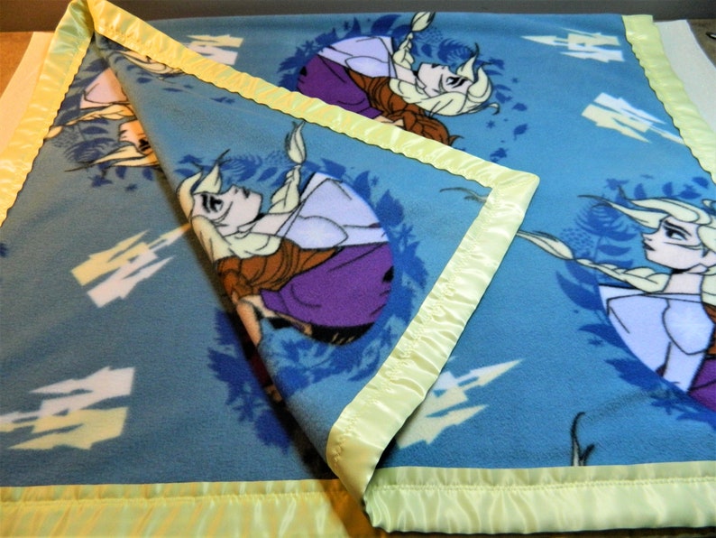 May include: A blue fleece blanket with a yellow satin border. The blanket features a repeated pattern of a cartoon image of a girl with long blonde hair.