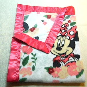 May include: A folded baby blanket featuring Minnie Mouse. The blanket has a white background with floral patterns in pink, peach, and green. It is trimmed with a bright pink satin border. Minnie Mouse is depicted in her classic outfit.