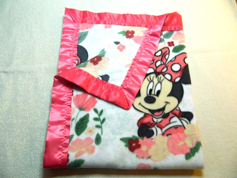 May include: A folded baby blanket featuring Minnie Mouse. The blanket has a white background with floral patterns in pink, peach, and green. It is trimmed with a bright pink satin border. Minnie Mouse is depicted in her classic outfit.