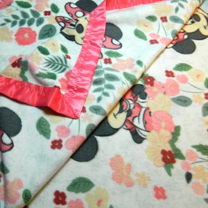 May include: A white blanket with a floral design and Minnie Mouse illustrations. The blanket has a pink satin border. The pattern includes pink, peach, and yellow flowers, and green leaves.