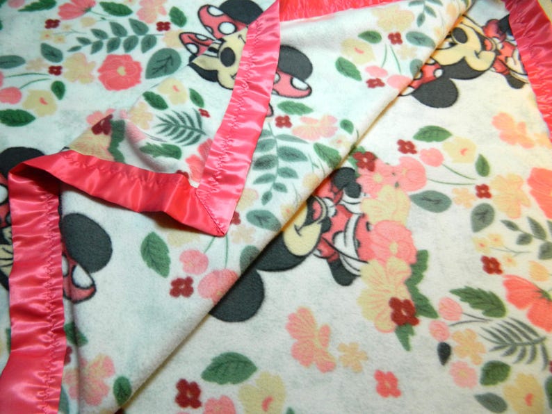 May include: A white blanket with a floral design and Minnie Mouse illustrations. The blanket has a pink satin border. The pattern includes pink, peach, and yellow flowers, and green leaves.