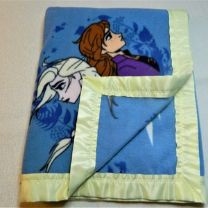 May include: A blue fleece blanket with a satin border. The blanket features a printed image of the characters Elsa and Anna from the movie Frozen.
