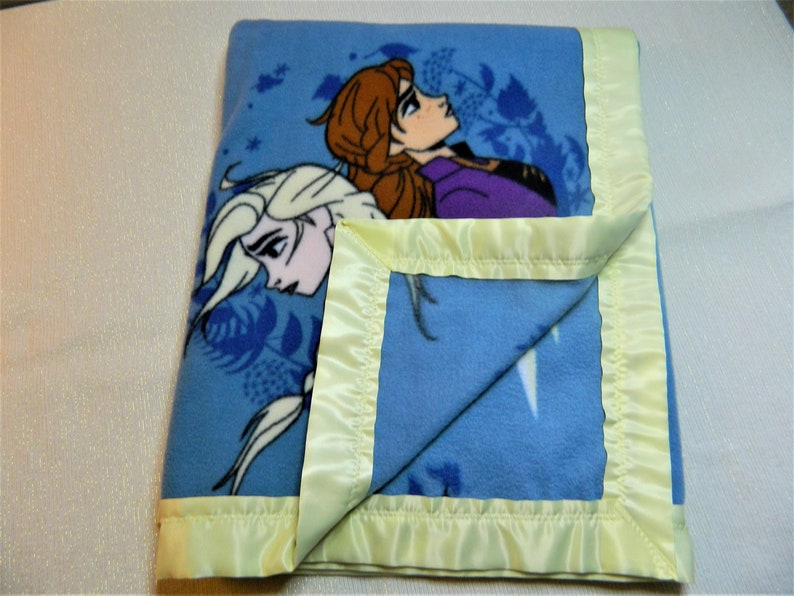 May include: A blue fleece blanket with a satin border. The blanket features a printed image of the characters Elsa and Anna from the movie Frozen.