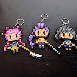 Pode incluir: Três chaveiros de pixel art com personagens em estilo anime com cabelos rosa, roxo e cinza. Cada chaveiro tem um chaveiro prateado e uma bengala de contas. Os personagens são feitos de contas de plástico coloridas.