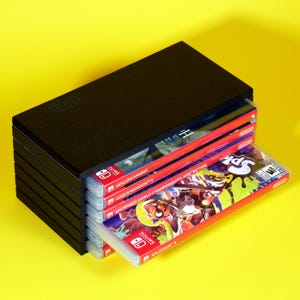以下が含まれることがあります： 黒いプラスチック製の収納ケースに、ニンテンドースイッチのゲームカートリッジが積み重ねられています。カートリッジには、「Hollow Knight」、「ルイージマンション3」、「Nintendo World Championships NES Edition」、「スプラトゥーン2」、「大乱闘スマッシュブラザーズ SPECIAL」のラベルが貼られています。