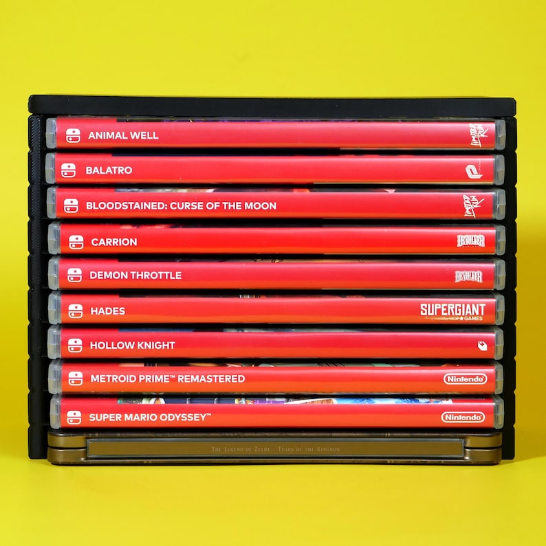 以下が含まれることがあります： ニンテンドースイッチ用のビデオゲームカートリッジ10本が積み重ねられています。カートリッジは縦に並べられており、各カートリッジにゲームタイトルが表示されています。タイトルには、「Animal Well」、「Balatro」、「Bloodstained: Curse of the Moon」、「Carrion」、「Demon Throttle」、「Hades」、「Hollow Knight」、「Metroid Prime Remastered」、「Super Mario Odyssey」が含まれています。