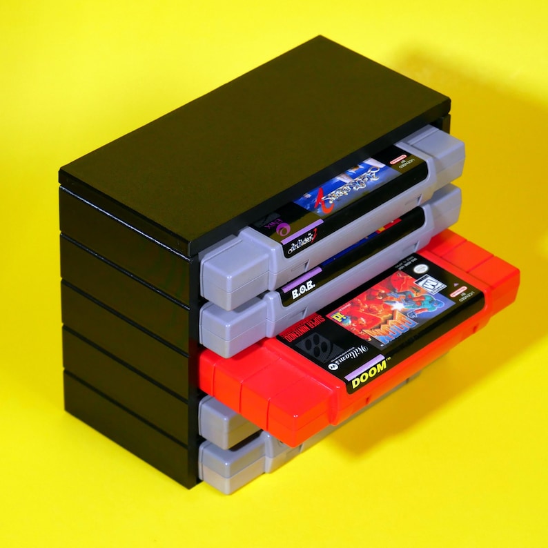 Puede incluir: Una caja de almacenamiento de pl&aacute;stico negro para cartuchos de juegos de Super Nintendo Entertainment System (SNES). La caja tiene capacidad para cinco cartuchos, con el cartucho superior parcialmente extra&iacute;do. El cartucho es rojo con el texto "DOOM" en &eacute;l. Los otros cartuchos son grises con varios t&iacute;tulos de juegos visibles.