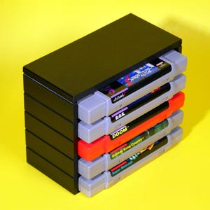 Puede incluir: Una caja de almacenamiento de pl&aacute;stico negro para cartuchos de juegos de Super Nintendo Entertainment System (SNES). La caja contiene cinco cartuchos, incluyendo juegos como "Doom", "Donkey Kong Country" y "Super Metroid".