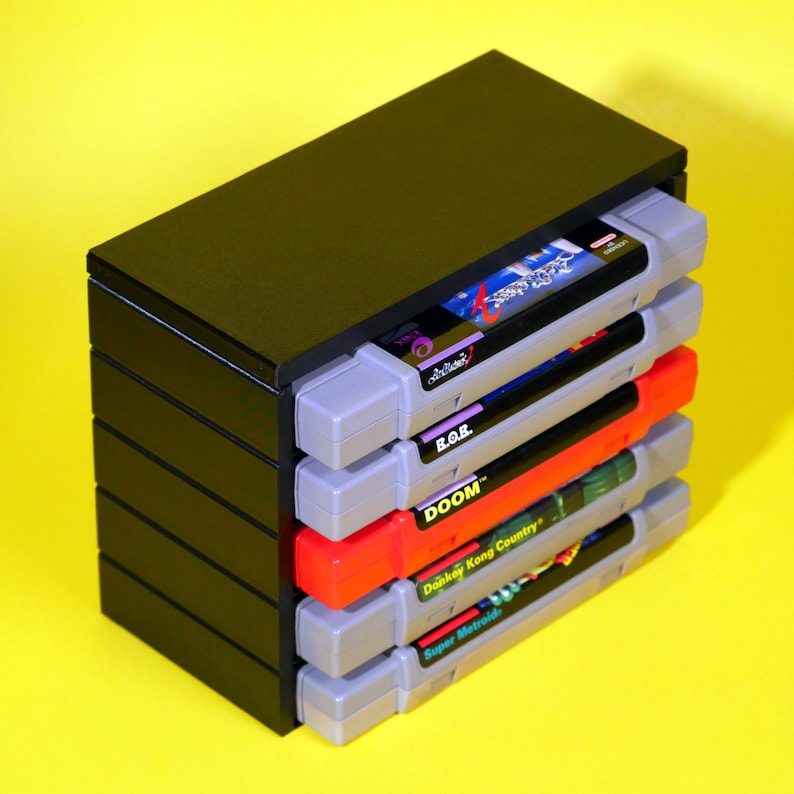 Puede incluir: Una caja de almacenamiento de pl&aacute;stico negro para cartuchos de juegos de Super Nintendo Entertainment System (SNES). La caja contiene cinco cartuchos, incluyendo juegos como "Doom", "Donkey Kong Country" y "Super Metroid".