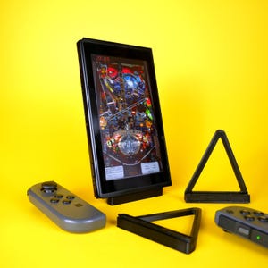 May include: A black Nintendo Switch console with a pinball game displayed on the screen. The console is on a stand and there are two black controllers and a black triangle stand next to it.