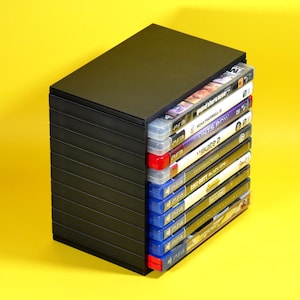 May include: A black plastic storage tower holding a vertical stack of ten video game cases. The collection includes various titles for PS3 and PS4 consoles, with the spines of the cases clearly visible, displaying game titles and console branding.
