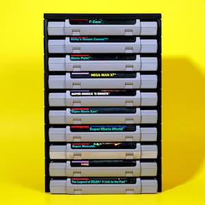 Puede incluir: Una pila de 10 cartuchos de juego de Super Nintendo Entertainment System (SNES) en una caja de almacenamiento de pl&aacute;stico gris. Los cartuchos est&aacute;n etiquetados con los nombres de juegos populares, incluyendo "F-Zero", "Kirby's Dream Course", "Mario Paint", "Mega Man X2", "Super Ghouls 'n Ghosts", "Super Mario Kart", "Super Mario World", "Super Metroid" y "The Legend of Zelda: A Link to the Past".