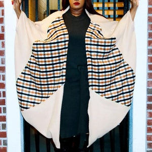May include: A beige wide-brimmed hat, a black ribbed mock-neck dress with a tie waist, and a beige open-front poncho featuring a large circular panel with a brown, black and white plaid pattern. The garment is paired with black knee-high boots.