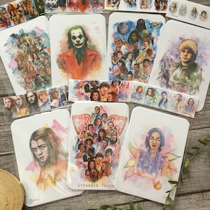 May include: A collection of watercolor paintings on white cardstock featuring various characters from popular movies and TV shows. The paintings include characters from Stranger Things, Star Wars, Joker, and more. The paintings are all in a similar style, with a watercolor background and a detailed portrait of the character.