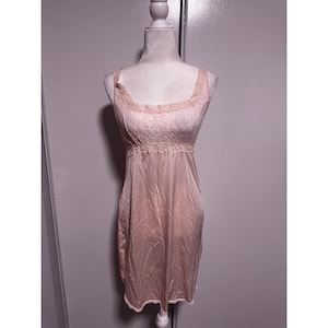 May include: A pale pink slip dress on a mannequin. The dress features a square neckline and straps with lace trim. The bodice has a textured pattern, and the waist is cinched with lace detailing. The skirt flows down to the knee.