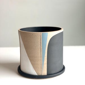 May include: A cylindrical ceramic planter with a black rim and base. The exterior features geometric shapes in white, tan, light blue, and dark gray. The planter is designed for indoor or outdoor use.