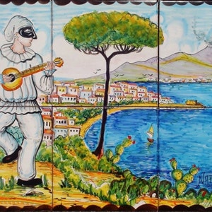 May include: A hand-painted ceramic tile depicting a man in a white costume playing a guitar, with a view of a town, a mountain, and a body of water in the background. The tile is decorated with a blue and green color scheme and features the word "NAPOLI" in the bottom right corner.