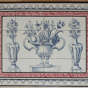 May include: A white ceramic tile with a blue and white floral design. The design features a large vase with flowers and fruit, flanked by two smaller vases with flowers. The tile is surrounded by a red and white border.