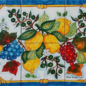 May include: A hand-painted ceramic tile set featuring a colorful still life of lemons, grapes, and leaves. The tile set is framed with a gold and blue border and includes the text "Dipinto a Mano Vietri".