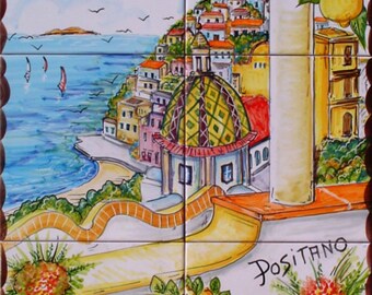May include: A colorful hand-painted ceramic tile mural depicting a view of Positano, Italy. The mural features a lemon tree, a colorful cityscape, a blue sea, and a white building with a green dome. The mural is framed by a white border and has the word "Positano" written in cursive at the bottom.