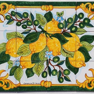 Pode incluir: Um mural de azulejos de cerâmica pintados à mão que representa um ramo de limoeiro com limões amarelos, folhas verdes e flores brancas. O azulejo é emoldurado por uma moldura verde e dourada e tem o texto "Ceramiche D'Italia" na parte inferior.