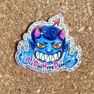 May include: A sticker featuring a blue cartoon demon with purple horns and a red arrow. The text "ALL DAY HYPE DEMON" is displayed around the demon. The sticker has a glittery border and the demon is giving the middle finger.