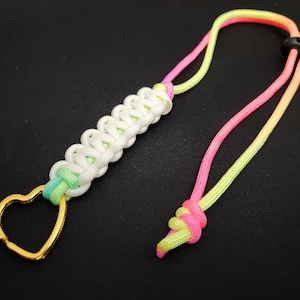 May include: A white, green, and pink paracord keychain with a gold heart-shaped ring. The keychain is made with a braided paracord and has a black plastic clasp.