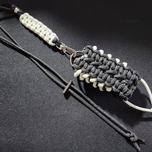 May include: A black and white paracord keychain with a silver clip and a small silver T-shaped charm. The keychain is made with a braided cord and has a white loop at the end.