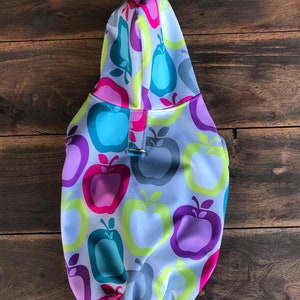 Może przedstawiać: Biała kurtka z kapturem dla psa z kolorowym nadrukiem jabłek. Kurtka ma sznurek u dołu i zamek błyskawiczny.