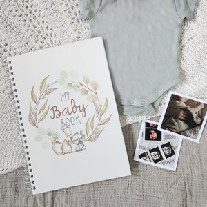 May include: A white baby book with a watercolor wreath of eucalyptus leaves and branches. The book has the words "My Baby Book" in a brown script font. A kangaroo and koala are holding hands inside the wreath. The book is lying on a white knitted blanket with a baby onesie and ultrasound photos.