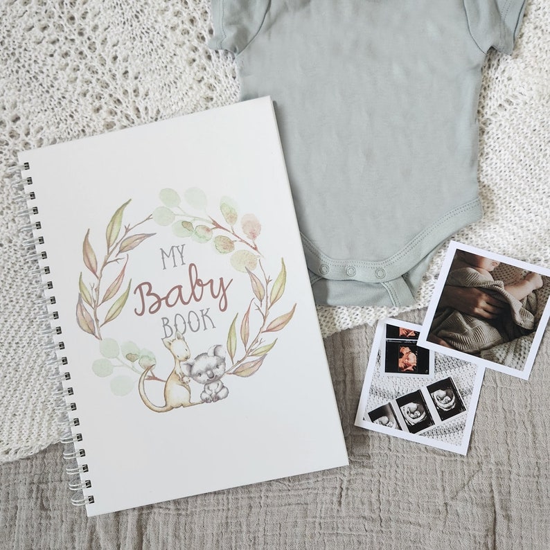 May include: A white baby book with a watercolor wreath of eucalyptus leaves and branches. The book has the words "My Baby Book" in a brown script font. A kangaroo and koala are holding hands inside the wreath. The book is lying on a white knitted blanket with a baby onesie and ultrasound photos.