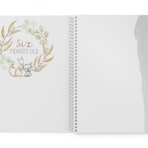 May include: A white spiral bound journal with a watercolor illustration of a koala and a kangaroo in a wreath of green leaves. The text "Six Months Old" is written in brown lettering.