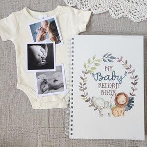 Puede incluir: Un libro blanco para registrar los recuerdos del beb&eacute; con un dise&ntilde;o de corona floral verde, marr&oacute;n y azul. La corona tiene un elefante y un le&oacute;n dentro. El texto "My Baby Record Book" est&aacute; en el centro de la corona. Hay tres fotos en blanco y negro en un body blanco.