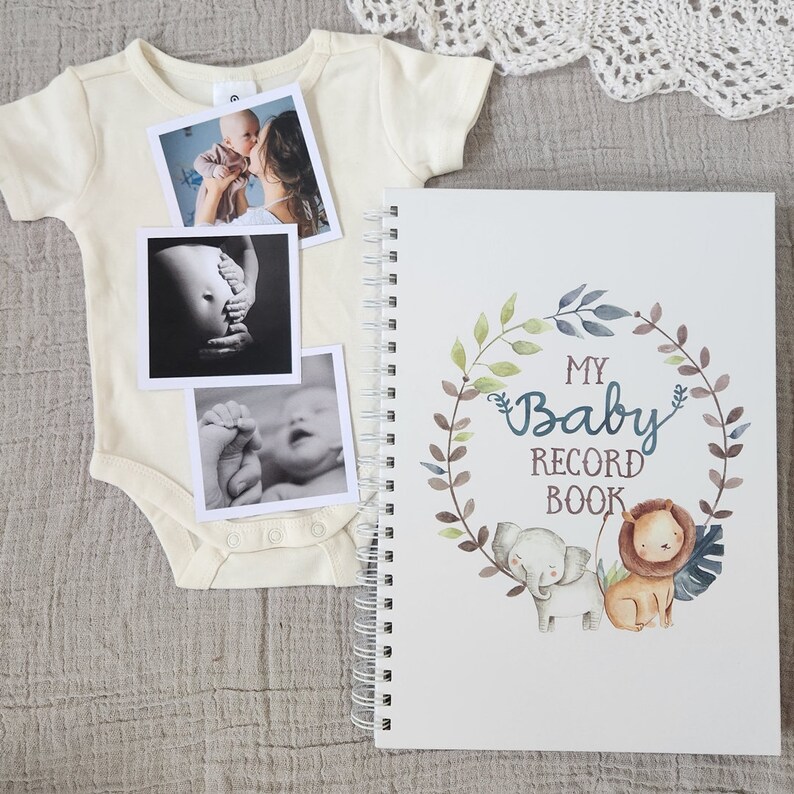 Puede incluir: Un libro blanco para registrar los recuerdos del beb&eacute; con un dise&ntilde;o de corona floral verde, marr&oacute;n y azul. La corona tiene un elefante y un le&oacute;n dentro. El texto "My Baby Record Book" est&aacute; en el centro de la corona. Hay tres fotos en blanco y negro en un body blanco.