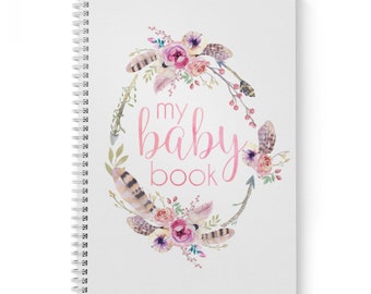 Puede incluir: Un libro de bebé con espiral y cubierta blanca. La portada presenta el texto "my baby book" en rosa, rodeado de una corona floral de acuarela con rosas rosas, plumas y vegetación. Para registrar los recuerdos del bebé.