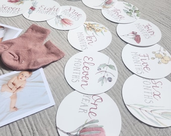 May include: A collection of baby milestone cards with floral illustrations and text. The cards are arranged around a pair of pink baby socks and a photo of a baby. The cards are white with floral designs and text such as "One Month" and "One Year".