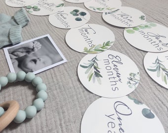 Può includere: Un set di biglietti per le tappe fondamentali del bambino con disegni di foglie di eucalipto ad acquerello. I biglietti sono bianchi con testo che indica i mesi e una foto del bambino. Sono visibili anche un anello per la dentizione verde menta e calzini per bambini.