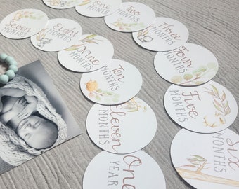 May include: A collection of round baby milestone cards with botanical illustrations and text. The cards are white with watercolor designs and text such as "hello world", "one month", and "one year". A black and white photo of a baby is also in the image.