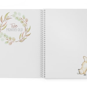 May include: A white spiral bound journal with a watercolor illustration of a wreath of leaves and the text "Two Months Old". On the right side of the journal is a watercolor illustration of a platypus.