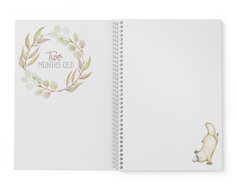 May include: A white spiral bound journal with a watercolor illustration of a wreath of leaves and the text "Two Months Old". On the right side of the journal is a watercolor illustration of a platypus.