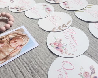 May include: A collection of round baby milestone cards with floral and feather designs, featuring months and "One year" in pink script. A photo of a sleeping baby in a knitted outfit and a pink teething ring are also present.