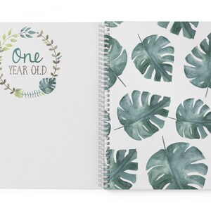 Puede incluir: Un cuaderno blanco de espiral con una corona de hojas verdes en la portada que dice "One Year Old". La contraportada tiene un patr&oacute;n repetitivo de hojas de monstera verdes en acuarela.