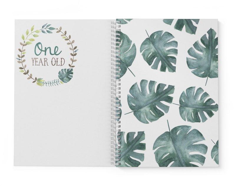 Puede incluir: Un cuaderno blanco de espiral con una corona de hojas verdes en la portada que dice "One Year Old". La contraportada tiene un patr&oacute;n repetitivo de hojas de monstera verdes en acuarela.