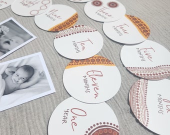 Può includere: Un set di 12 carte rotonde per i traguardi del bambino con sfondo bianco e bordi con motivi rossi e arancioni. Le carte presentano i numeri da uno a dodici, con la parola "mesi" scritta sotto ogni numero. La carta per un anno ha la parola "anno" scritta sotto il numero uno.