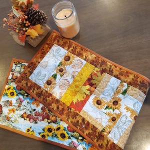 May include: Two rectangular quilted fabric placemats featuring autumn-themed patterns. One displays a vibrant scene of sunflowers, pumpkins, and rustic buildings, while the other features vertical strips of sunflowers, leaves, and pumpkins in orange, yellow, and white.