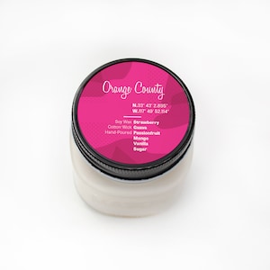 Puede incluir: Un tarro de vela rosa y blanco con una tapa negra. La etiqueta dice "Orange County" con coordenadas y una lista de aromas: Fresa, Guava, Maracuy&aacute;, Mango, Vainilla, Az&uacute;car. La vela est&aacute; hecha con cera de soja y una mecha de algod&oacute;n.