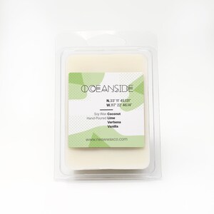 May include: A white soy wax candle in a clear plastic container. The label is green and white with the text "OCEANSIDE", "N.33° 11' 45.131", "W.117° 22' 46.14", "Soy Wax Hand-Poured", "Coconut Lime Verbena Vanilla", and "www.neswwaxco.com".